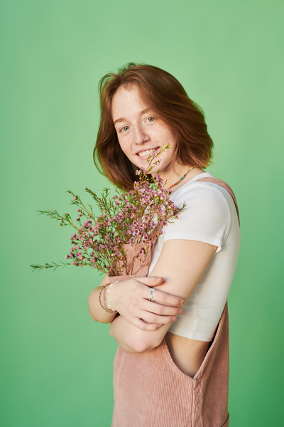 A woman with red hair dressed in a pink corduroy sundress decorated with pink wildflowers poses against a green background