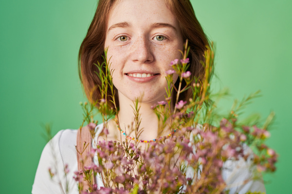 Close-up of a smiling woman with short red hair with a bouquet of pink and yellow wildflowers against a green background