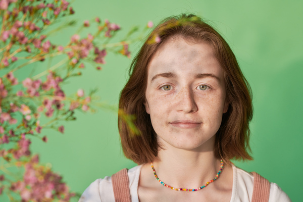 A portrait of a young woman with short red hair and freckles with a colored beaded necklace framed by pink wildflowers