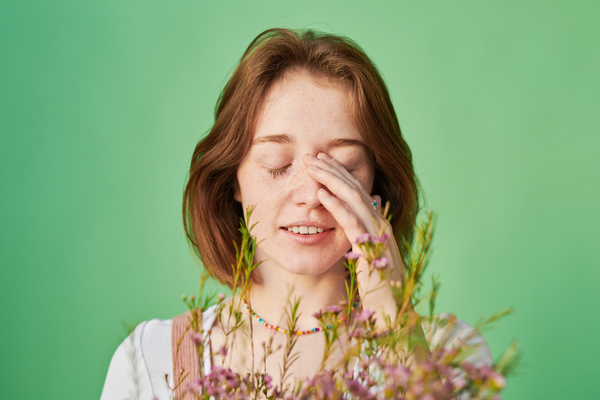 Close-up of a woman with short red hair with a bouquet of pink and yellow wildflowers against a green background closing her eyes and touching her face
