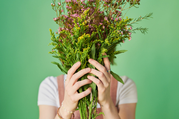 Close-up of a woman hiding her face behind a bouquet of pink and yellow wildflowers stood against a green background