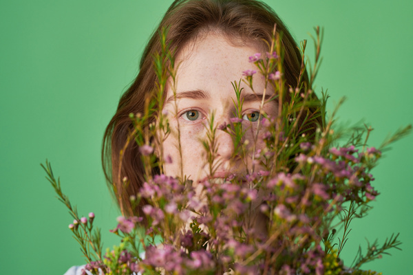 The face of a woman with red hair and freckles behind a bouquet of pink wildflowers