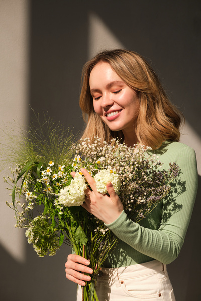 A smiling woman with blonde hair in light clothes with a large bouquet of flowers in a sun room