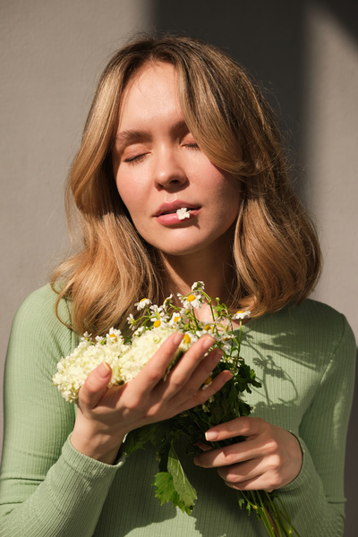 A woman with short blonde hair and a white flower bud on her lips posing witha bouquet of daisies with her eyes closed