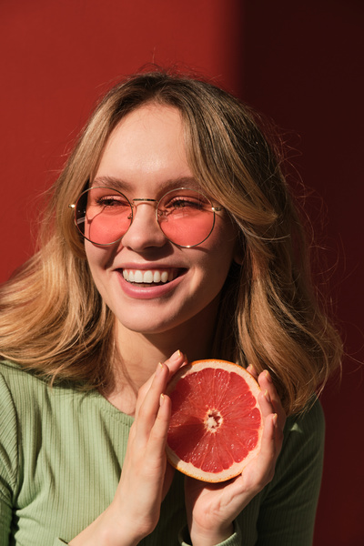 A laughing woman with light hair in a mint-colored longsleeve and sunglasses holding a half a grapefruit in her hands
