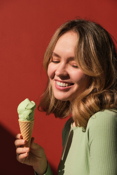 A woman with blonde hair posing against a red background with a pistachio ice cream in a waffle cone