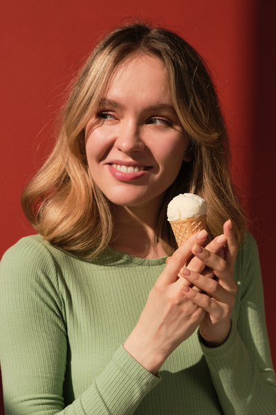 A woman with short blonde hair dressed in a green longsleeve looking to the side poses with vanilla ice cream in a cone against a red background
