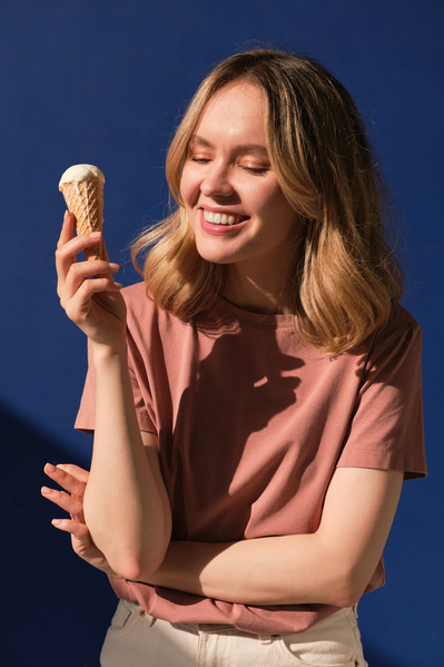 A smiling woman with short blonde hair wearing a pink T-shirt broadly holding a vanilla ice cream cone against a blue background