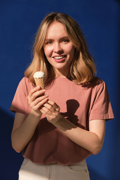 A woman with short blonde hair in a pink T-shirt and white jeans holding a melting vanilla ice cream in a cone against a blue background