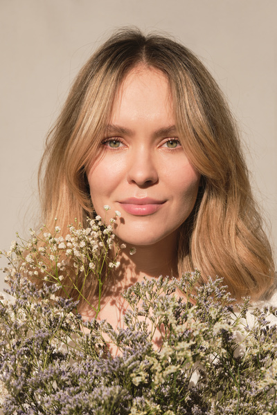 A blonde haired woman with gray eyes with a bouquet of wildflowers at her face against a white background