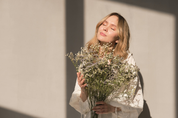 A blonde haired woman with closed eyes posing with a bouquet of wildflowers against a white wall in a sunny room