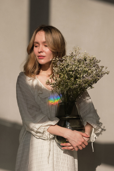 A woman with blonde hair dressed in a white dress holding a dark vase with a bouquet of light wildflowers
