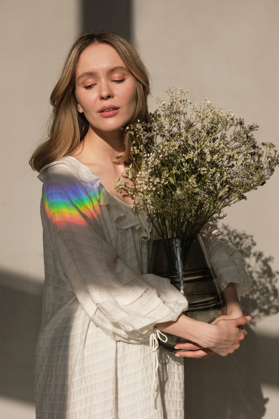 A woman with blonde hair dressed in a white dress posing with a dark vase of light wildflowers and a rainbow ray on her shoulder