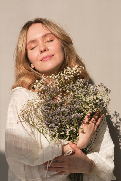 A young blonde woman in a light white outfit posing with a bouquet of small wildflowers of light shades