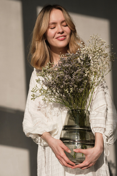 A blonde in a white robe posing with a black glass vase with a bouquet of small flowers of light shades in a sunny room