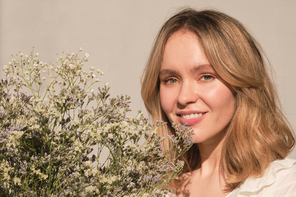 A young blonde woman with a bouquet of wildflowers of light shades near her face against a white background