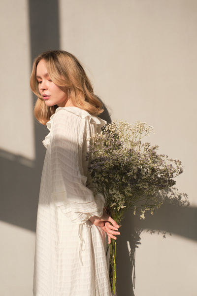 A young blonde woman in a white summer dress holding behind her back a bouquet of wildflowers of light shades against a white background