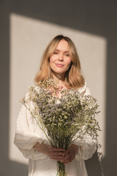 A blonde woman in a white dress with a bouquet of wildflowers stands against a white background