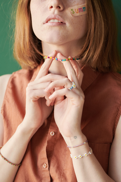 A red-haired woman in a red sleeveless blouse touching a multicolored beaded necklace with her hands with bracelets