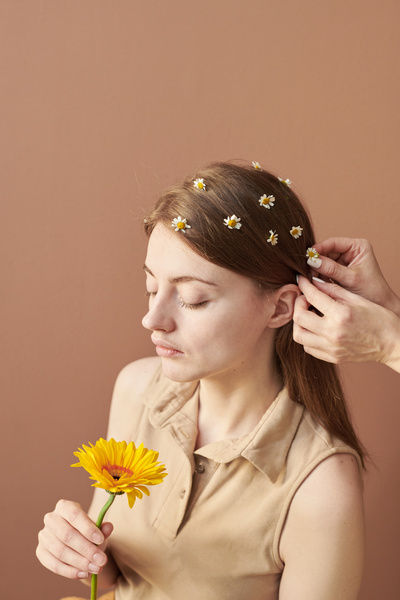 The hair of a young woman holding a yellow gerbera is adorned with camomiles against a beige background