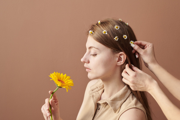 The hair of a beautiful woman with a yellow gerbera is adorned with daisies against a brown background