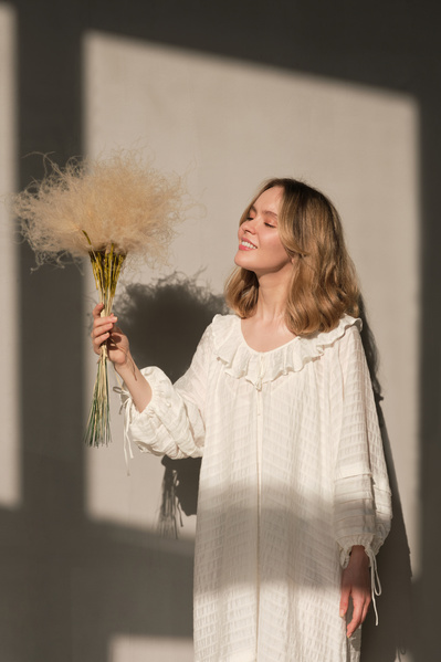 A blonde woman in a white light dress with dried flowers in her hand stands in a bright room