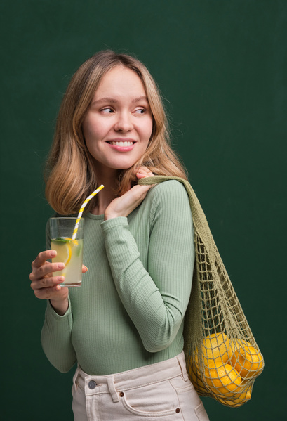 A blonde-haired woman dressed in summer clothes posing with a green organic string bag filled with citrus fruits and a refreshing drink