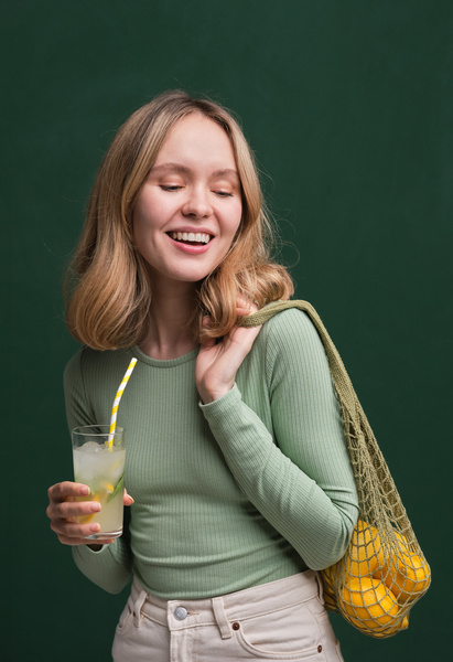 A woman with blonde hair dressed in summer clothes posing with citrus fruits in a green organic string bag and a refreshing drink
