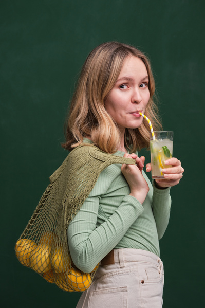A woman with blonde hair dressed in light clothes with citrus fruits in a green organic string bag drinking a summer lemonade