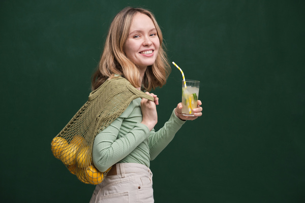 A blonde woman dressed in light clothes with citrus fruits in a green cotton string bag and a summer drink
