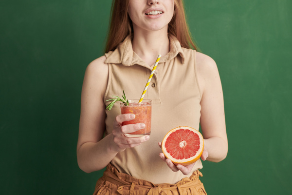 A woman dressed in beige clothes with a glass with a yellow cocktail straw full of a refreshing drink and half a grapefruit in the hands
