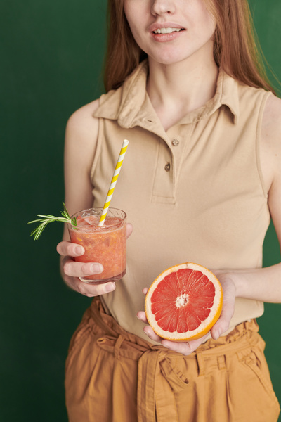 A glass with a yellow cocktail straw filled with a summer drink and half a grapefruit in the hands of a female dressed in light clothes
