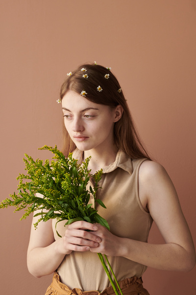A young woman with brown hair adorned with camomiles posing with wildflowers against a brown background