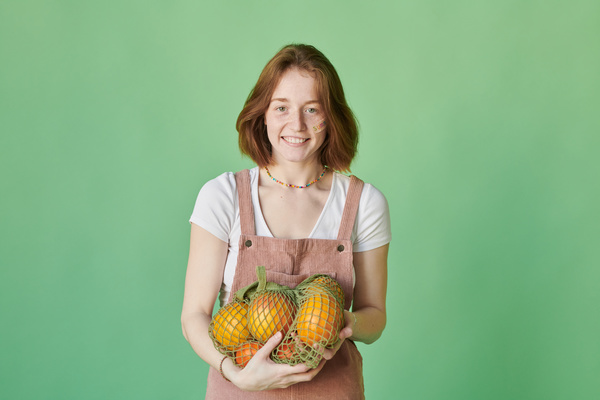 A woman with short red hair dressed in summer dress posing with a green organic string bag with citrus fruits