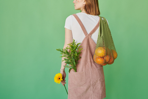 A woman with red hair dressed in a corduroy sundress and standing with her back having flowers and citrus fruits in a green cotton string bag