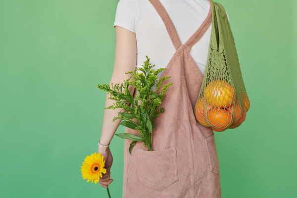 A woman with red hair a corduroy sundress with flowers in the pocket holding a green organic string bag with oranges