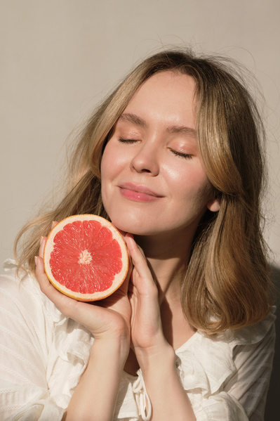 A blonde woman with closed eyes in a white dress posing with half a grapefruit against a white wall