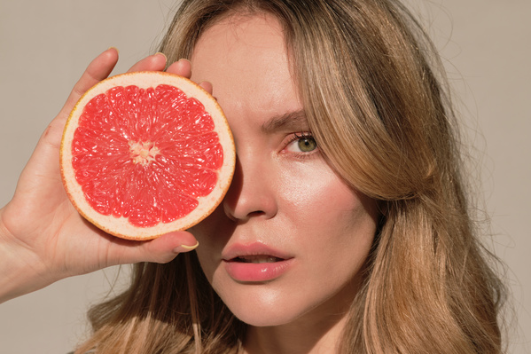 A blonde woman in a white dress posing with half a grapefruit hiding her eye against a white wall