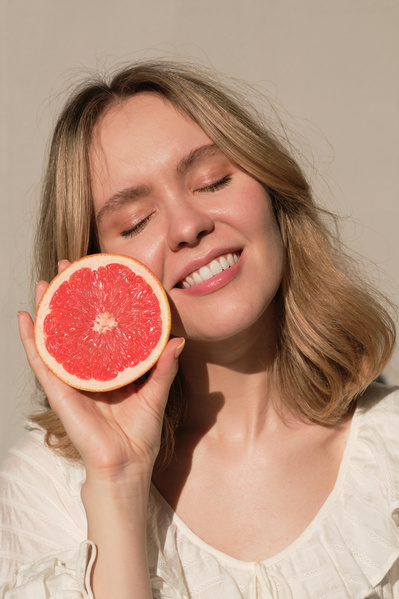 A smiling woman with white hair and closed eyes in a white dress holding half of a grapefruit at her face against a white wall
