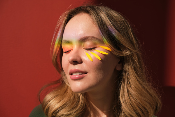 A woman with blonde hair and an rainbow ray and petals on her face against a red background