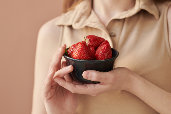 Close-up of a black bowl with strawberries in the hands of a woman wearing a beige singlet with a collar