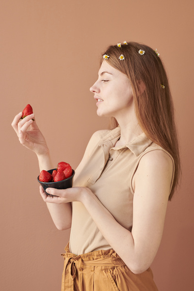 Profile of a woman with long hair adorned with chamomile buds looking at strawberry from a black bowl in her hand