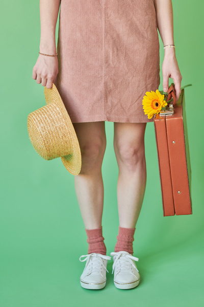 A woman in a corduroy pink sundress and white sneakers holding suitcase with a yellow gerbera and a straw hat in the hands