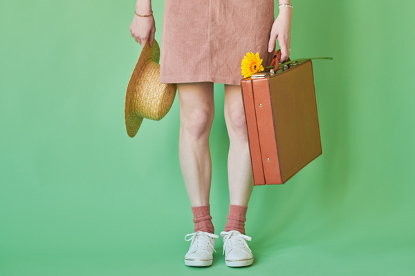 A woman in a corduroy pink sundress and white sneakers holding a suitcase with a yellow gerbera and a straw hat