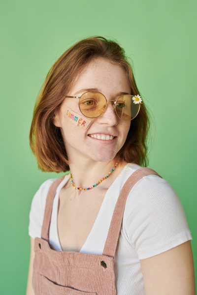 A red-haired woman with a sticker on her face wearing sunglasses with a daisy dressed in summer clothes against a green background