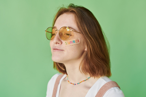 A redheaded woman with a summer sticker on her face wearing yellow sunglasses decorated with a daisy against a green background