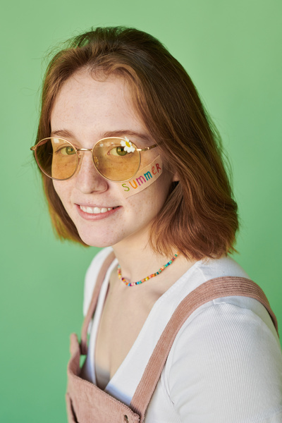 A red-haired woman with a summer decal on her face in yellow sunglasses with a camomile against a green background