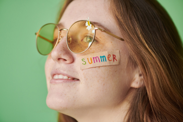 Summer sticker on the cheek of a woman with red hair in yellow sunglasses with a daisy against a green background
