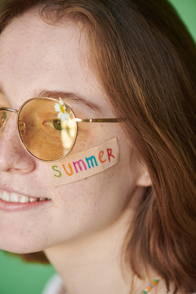 Summer decal on the cheek of a woman with red hair in yellow sunglasses with a camomile against a green background