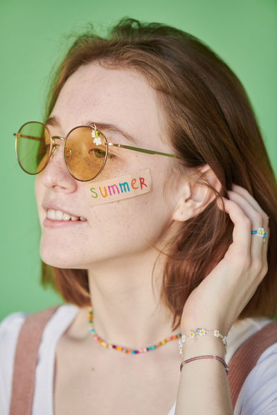 A woman in yellow sunglasses with a daisy and a sticker with the inscription on her face correcting her red hair